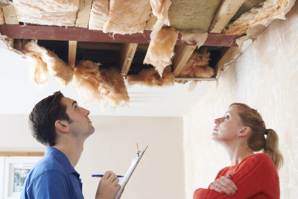 male & female standing in room with a hole in the ceiling, staring at the hole, the man is holding a clipboard